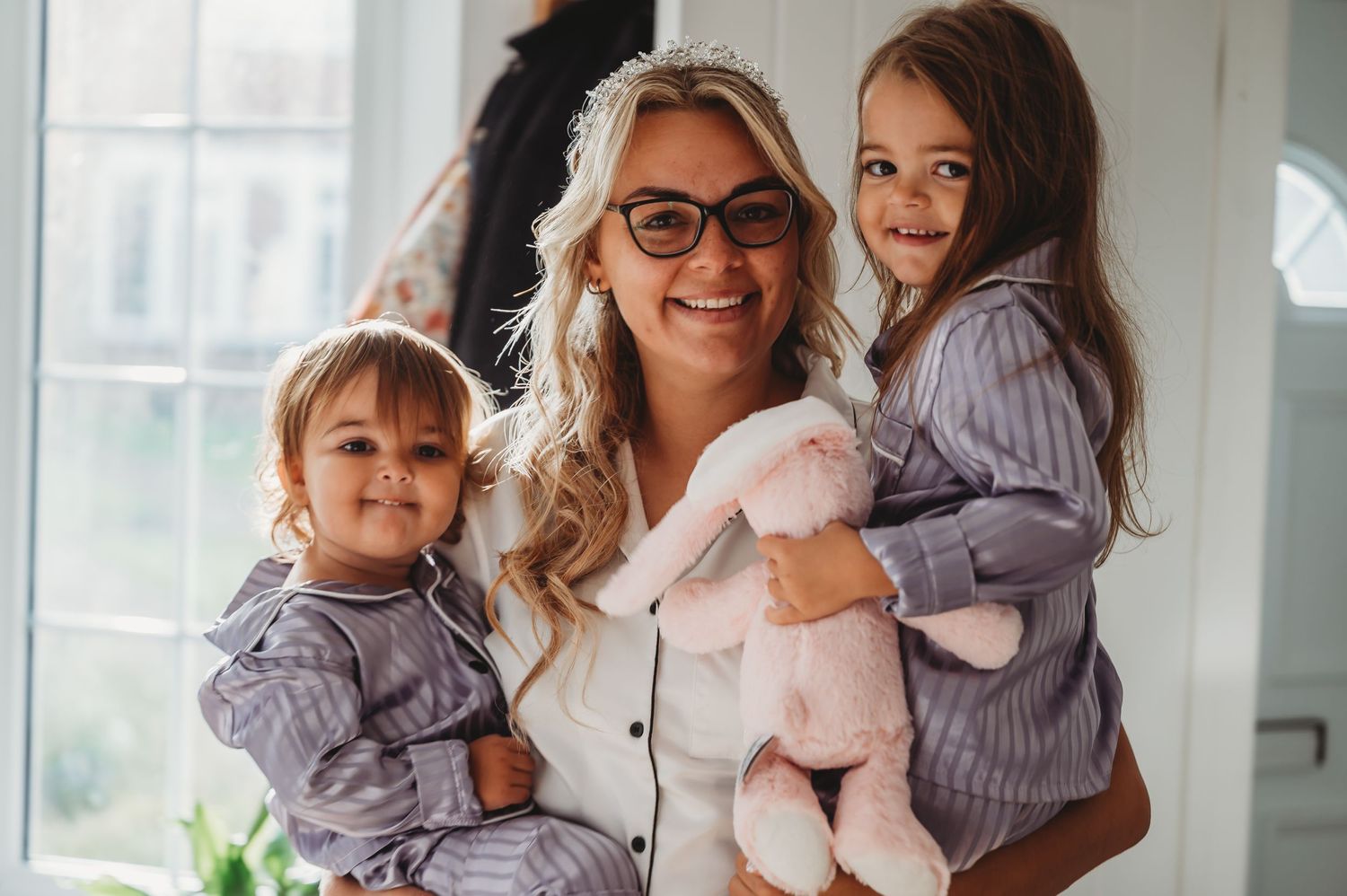 Three people wearing matching purple striped pajamas smile together while holding a pink plush toy.