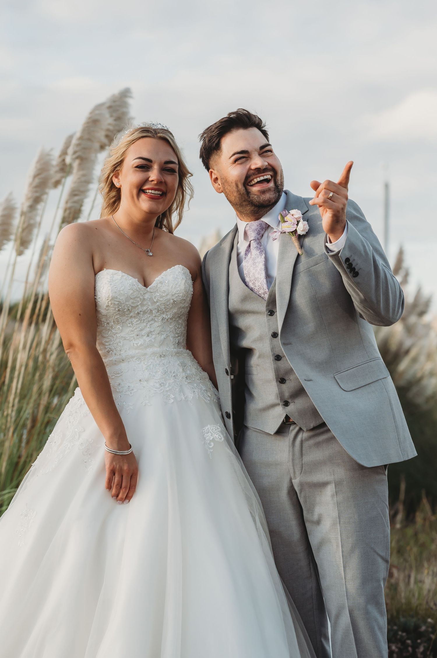 A joyful wedding couple poses outdoors among tall pampas grass, with the bride in white and groom in light gray suit.