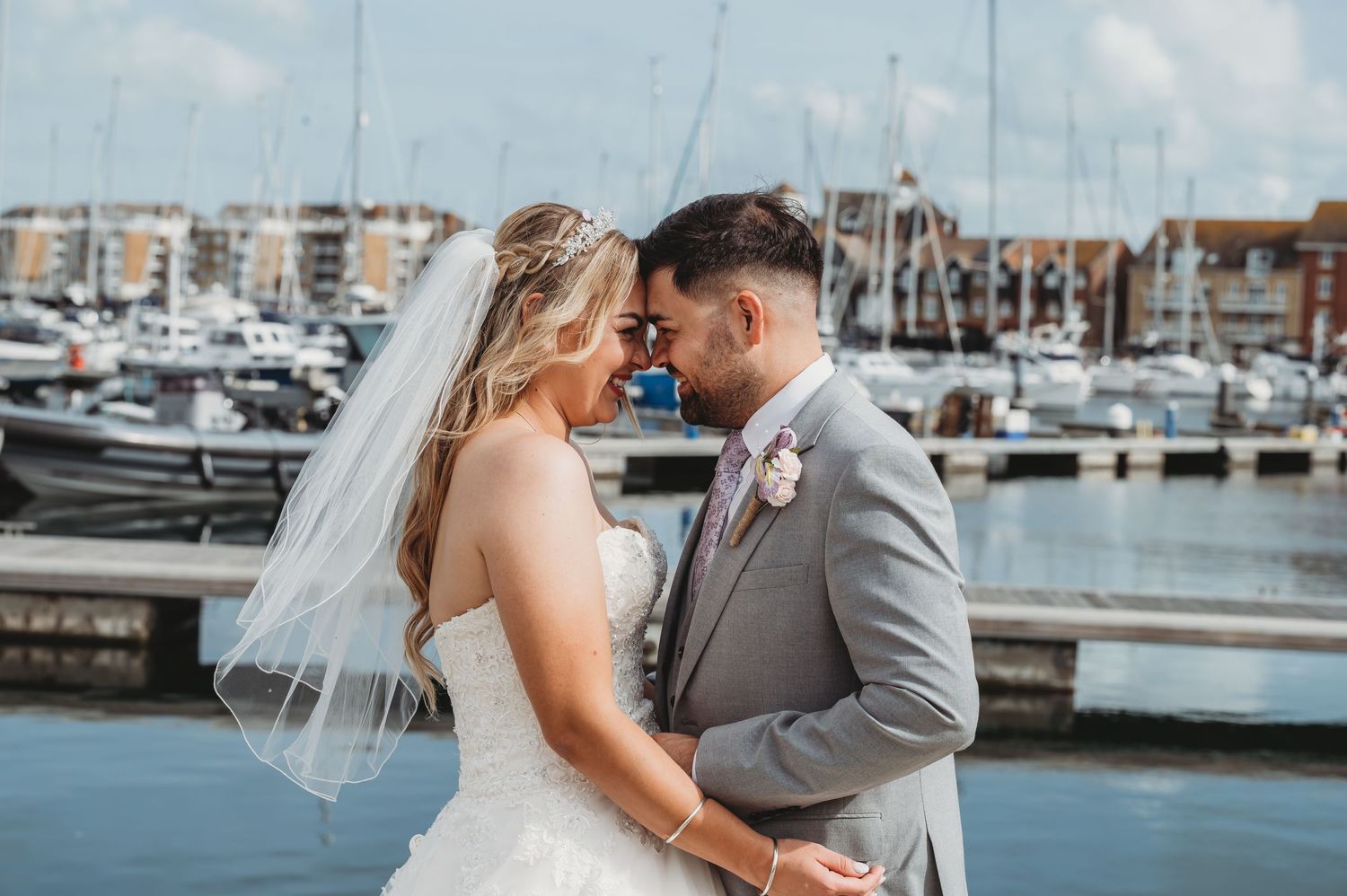 A romantic wedding couple embraces at a marina with boats and harbor buildings in the background.