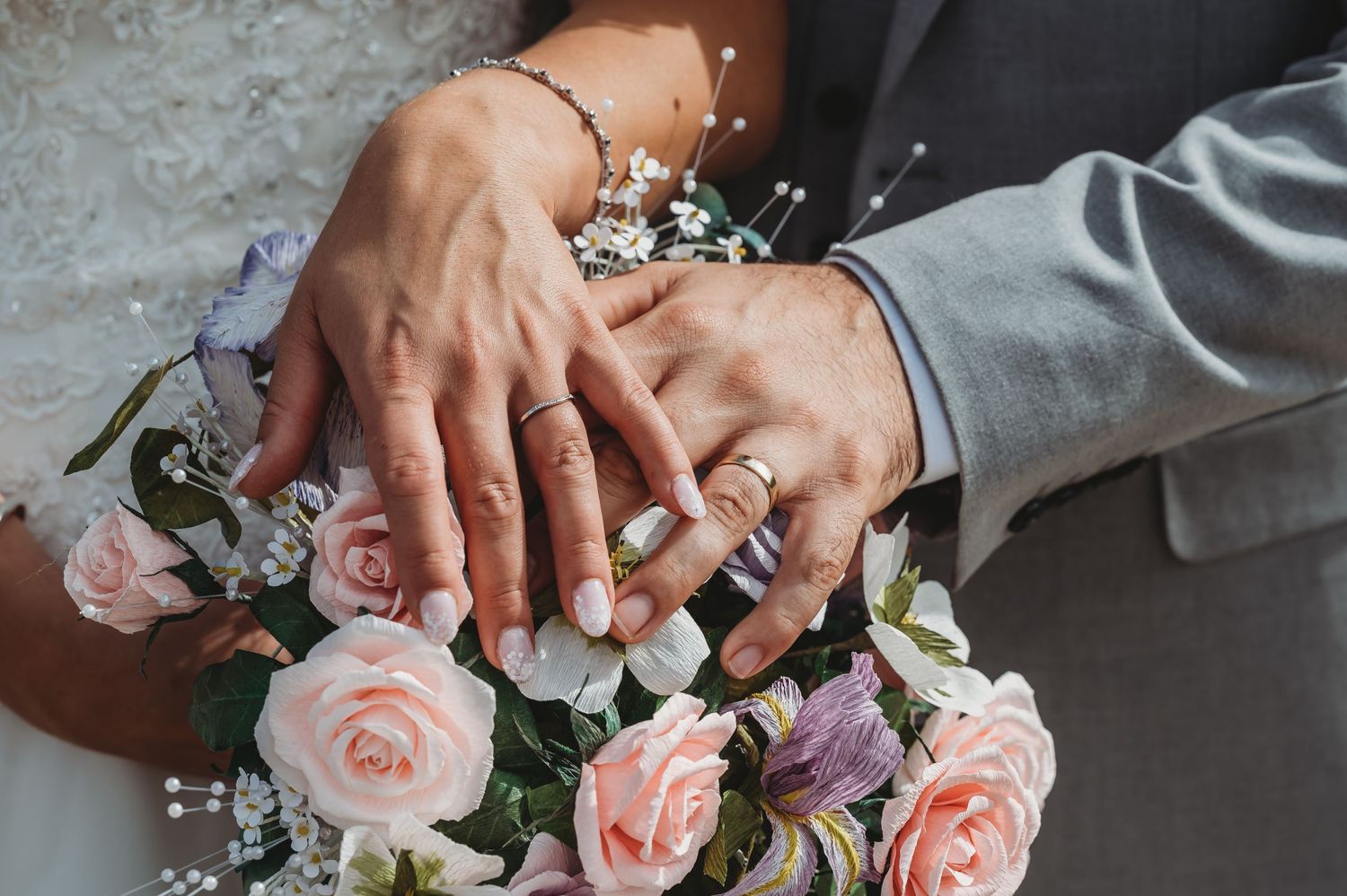 Hands with wedding rings resting on pink rose and baby's breath bouquet showing newlywed couple's jewelry.