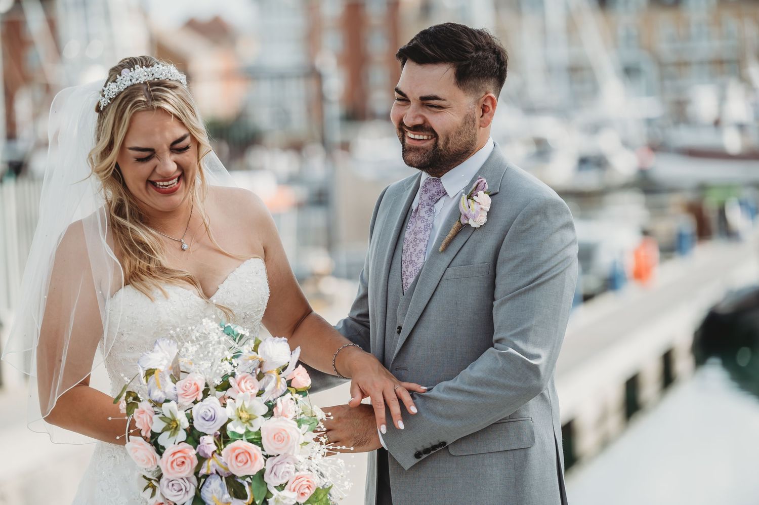 A joyful couple shares a candid moment at a marina dock, with the bride holding a pink and white floral bouquet.