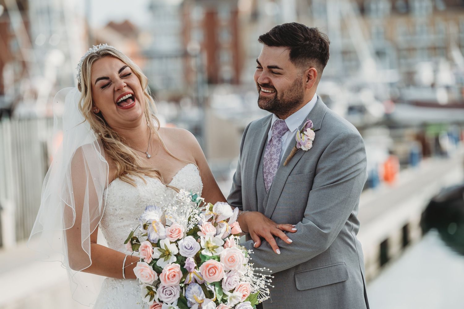 A joyful couple in wedding attire share a candid moment at a marina, with the bride holding a pink and white bouquet.