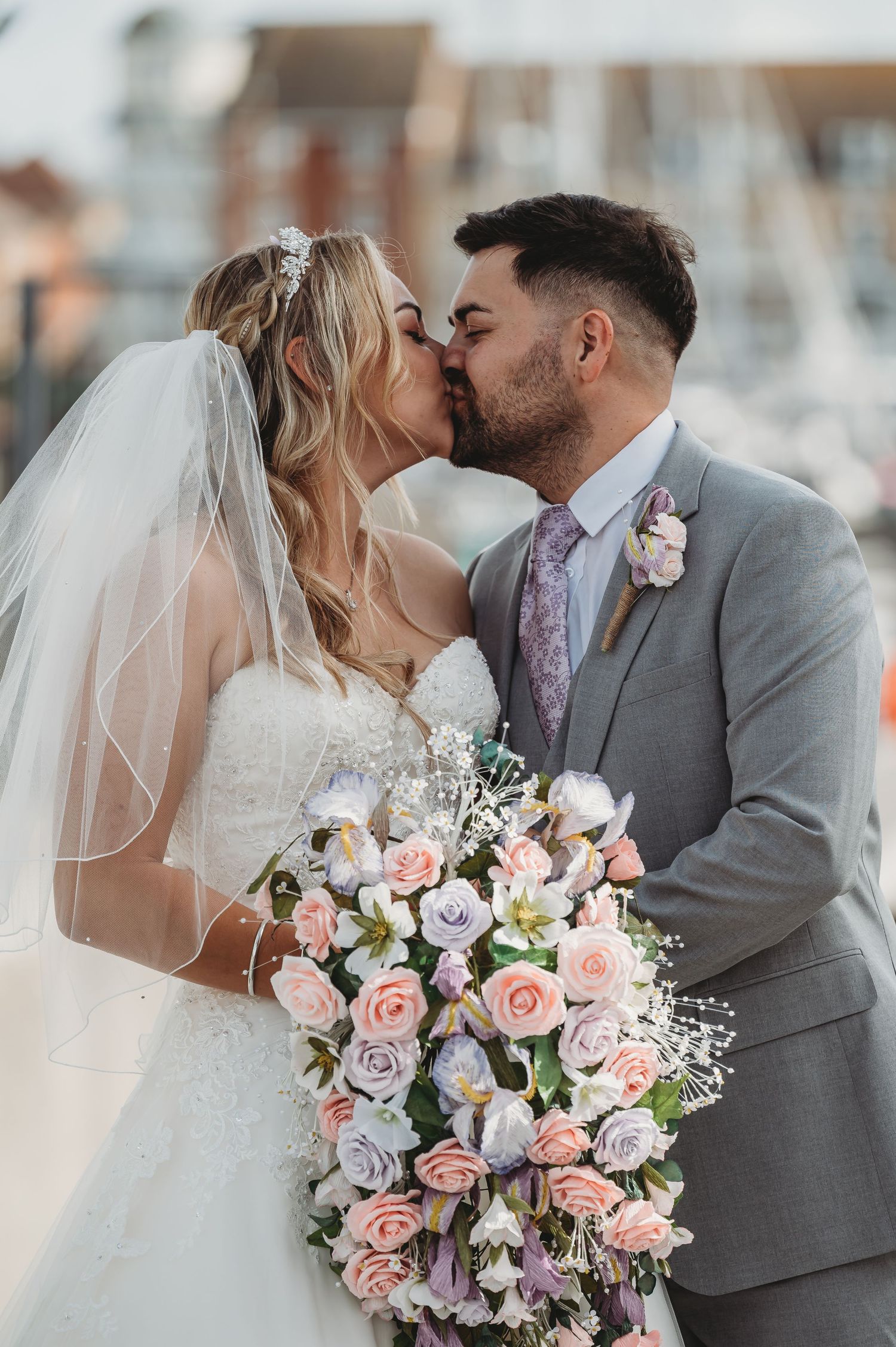 A wedding couple shares a romantic kiss while the bride holds a stunning pink and purple floral bouquet.