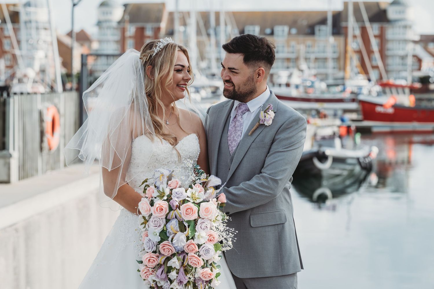 Newlyweds in wedding attire embrace at a marina dock with boats and pink flower bouquet in romantic coastal setting.