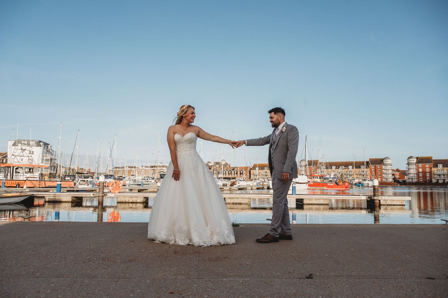 A couple celebrates their wedding day on a dock overlooking a scenic marina with boats in the background.