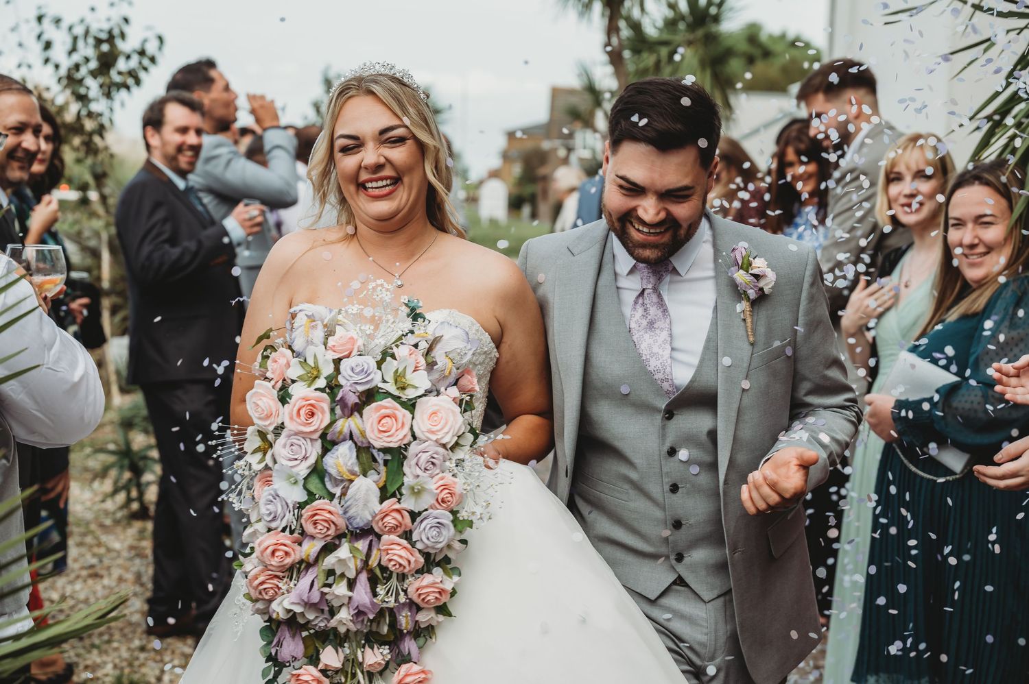 Joyful newlyweds walk together among celebrating guests while confetti falls at an outdoor wedding ceremony.