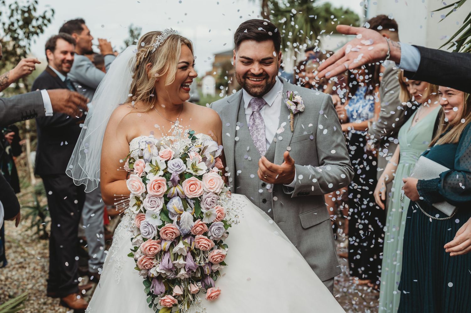 A joyful wedding couple walks through a shower of confetti while holding a pink and white rose bouquet.