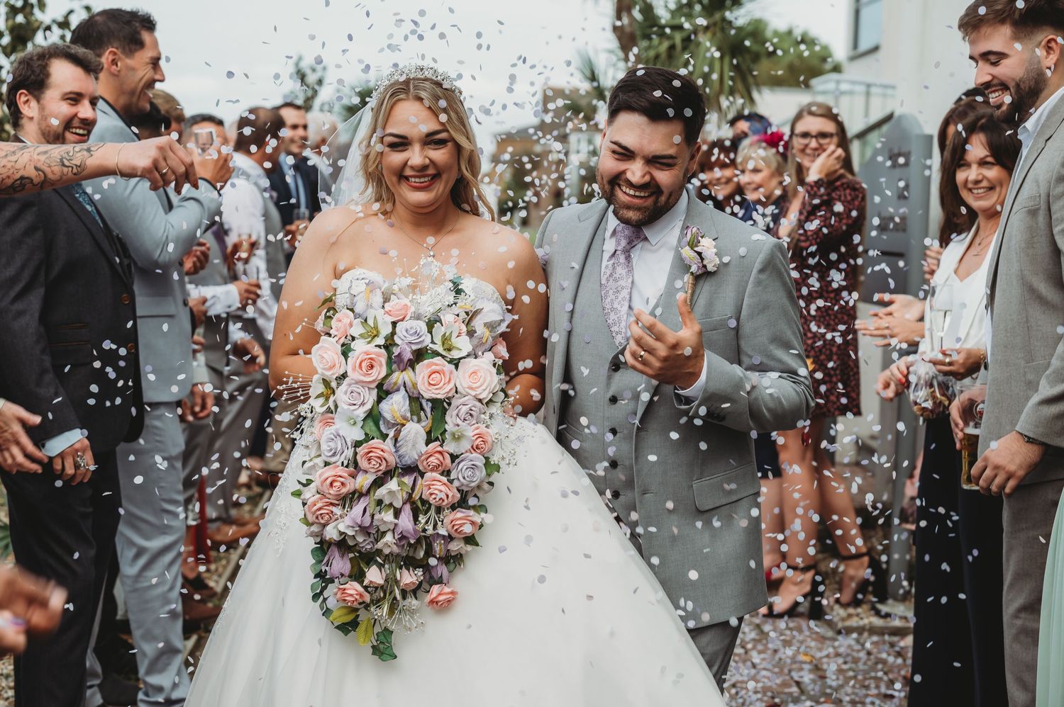 A joyful newlywed couple celebrates amid a shower of confetti while holding a colorful floral bouquet.