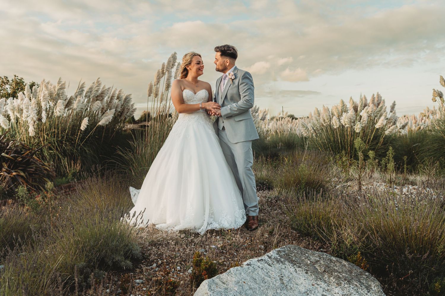 A couple in wedding attire poses together at sunset among tall grasses and a rocky landscape.