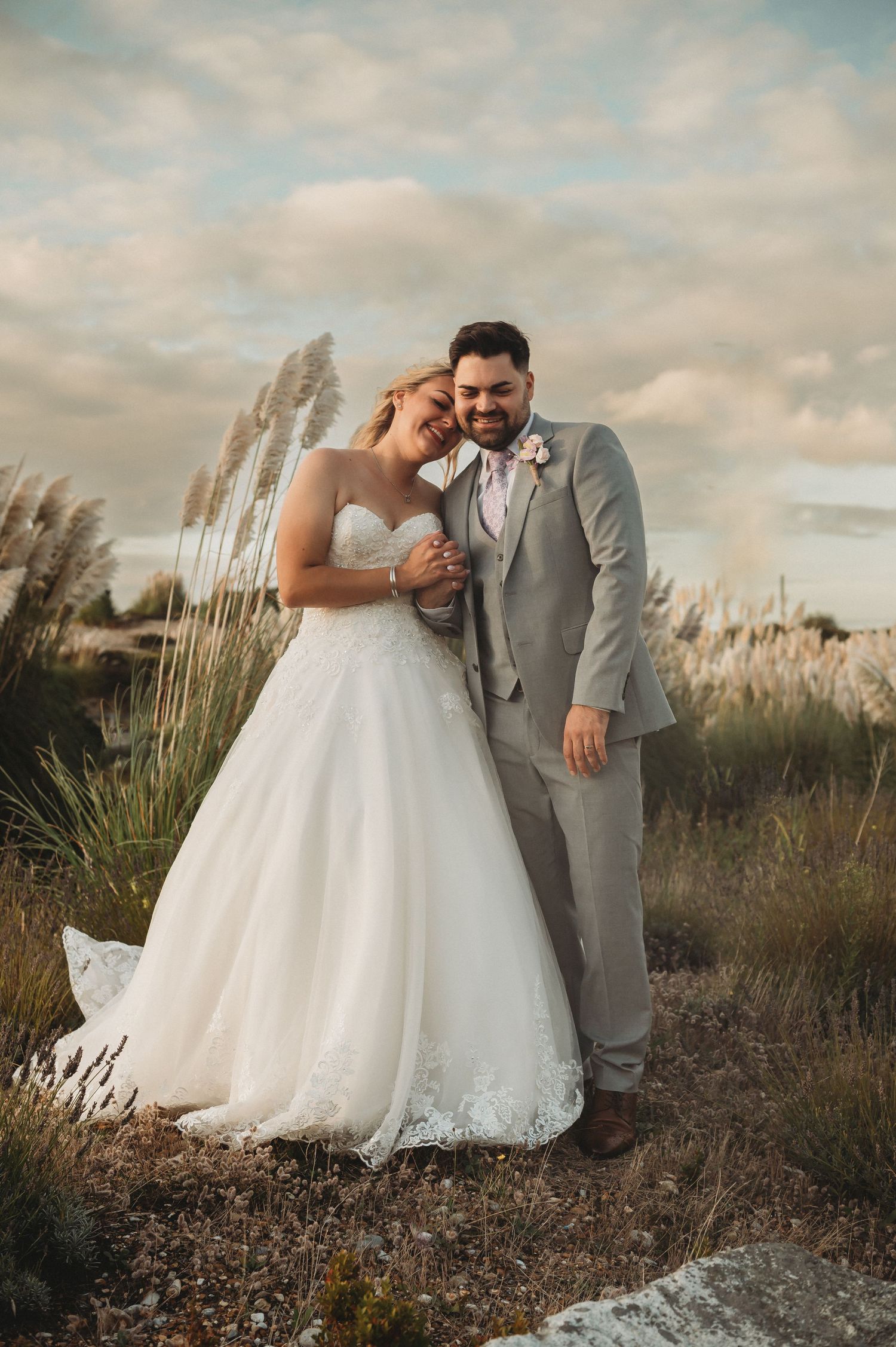 A romantic wedding photo of a couple embracing outdoors at sunset with pampas grass swaying in the background.