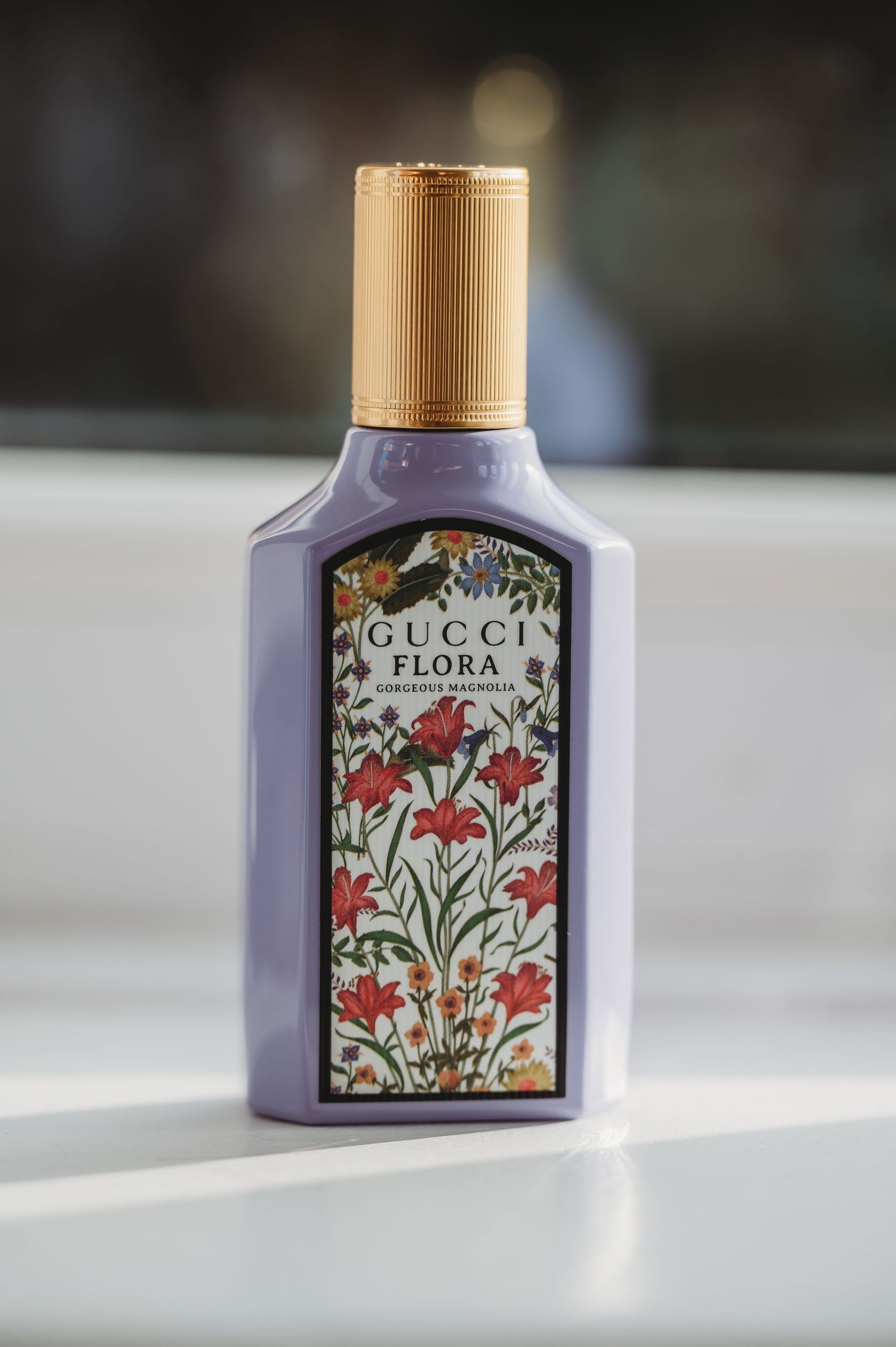 A bottle of Gucci Flora perfume with purple liquid and floral design on its label against a light background.