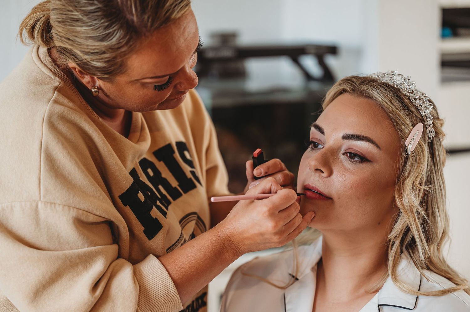 Makeup artist applying lipstick to client during professional beauty session while wearing tan sweatshirt.