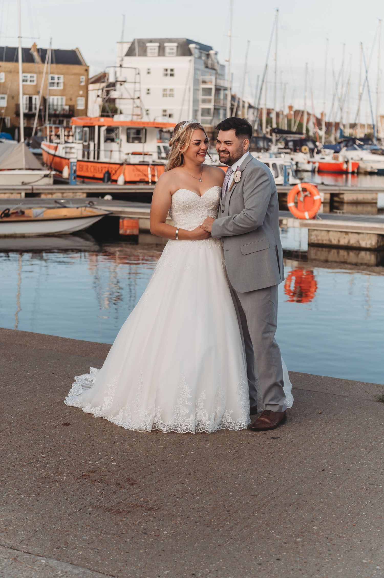 A wedding couple poses together at a marina dock with boats and orange buoys in the background at sunset.