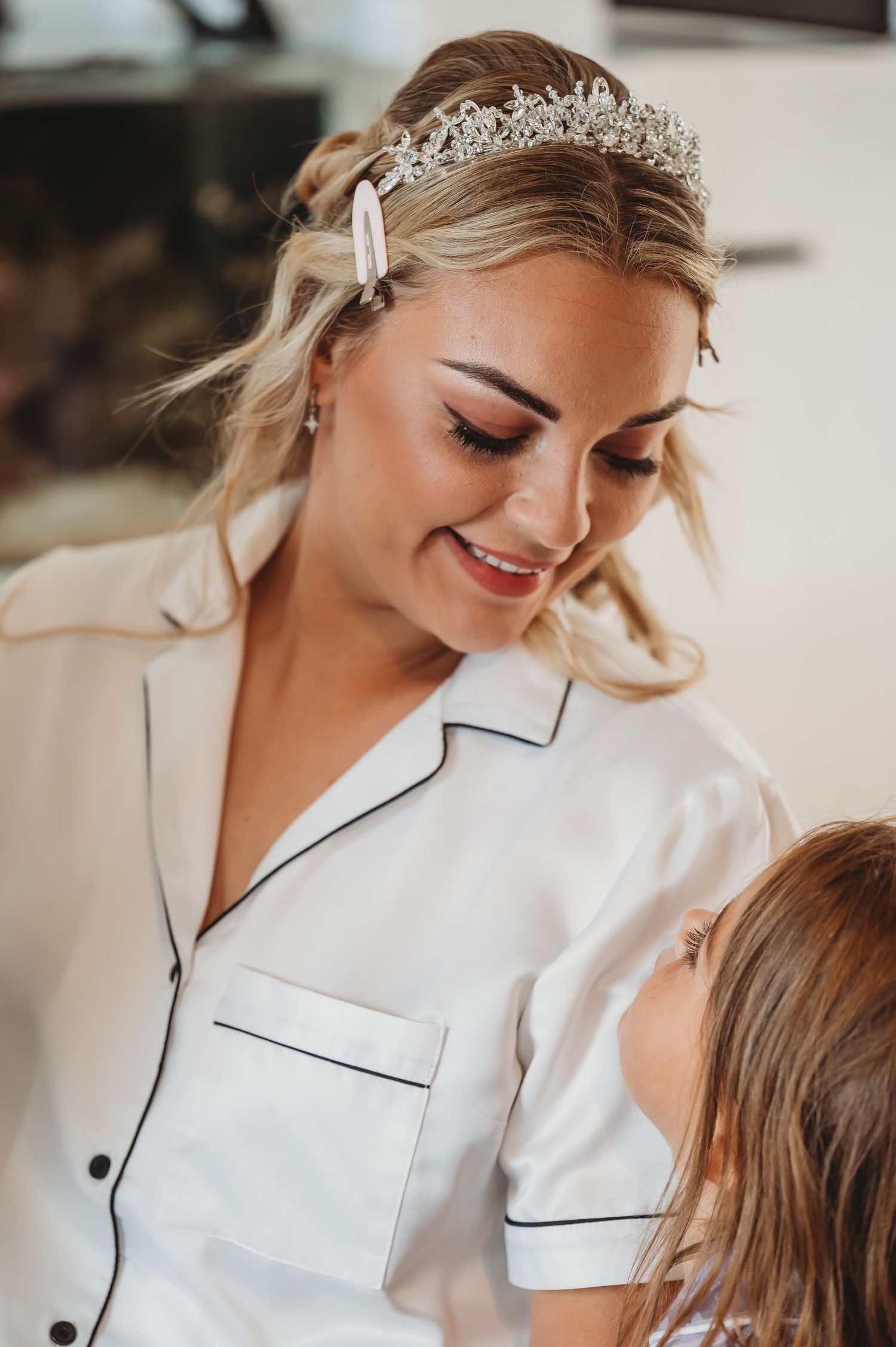 A bride wearing a white silk pajama top and crystal tiara looks down with a warm smile during wedding preparations.