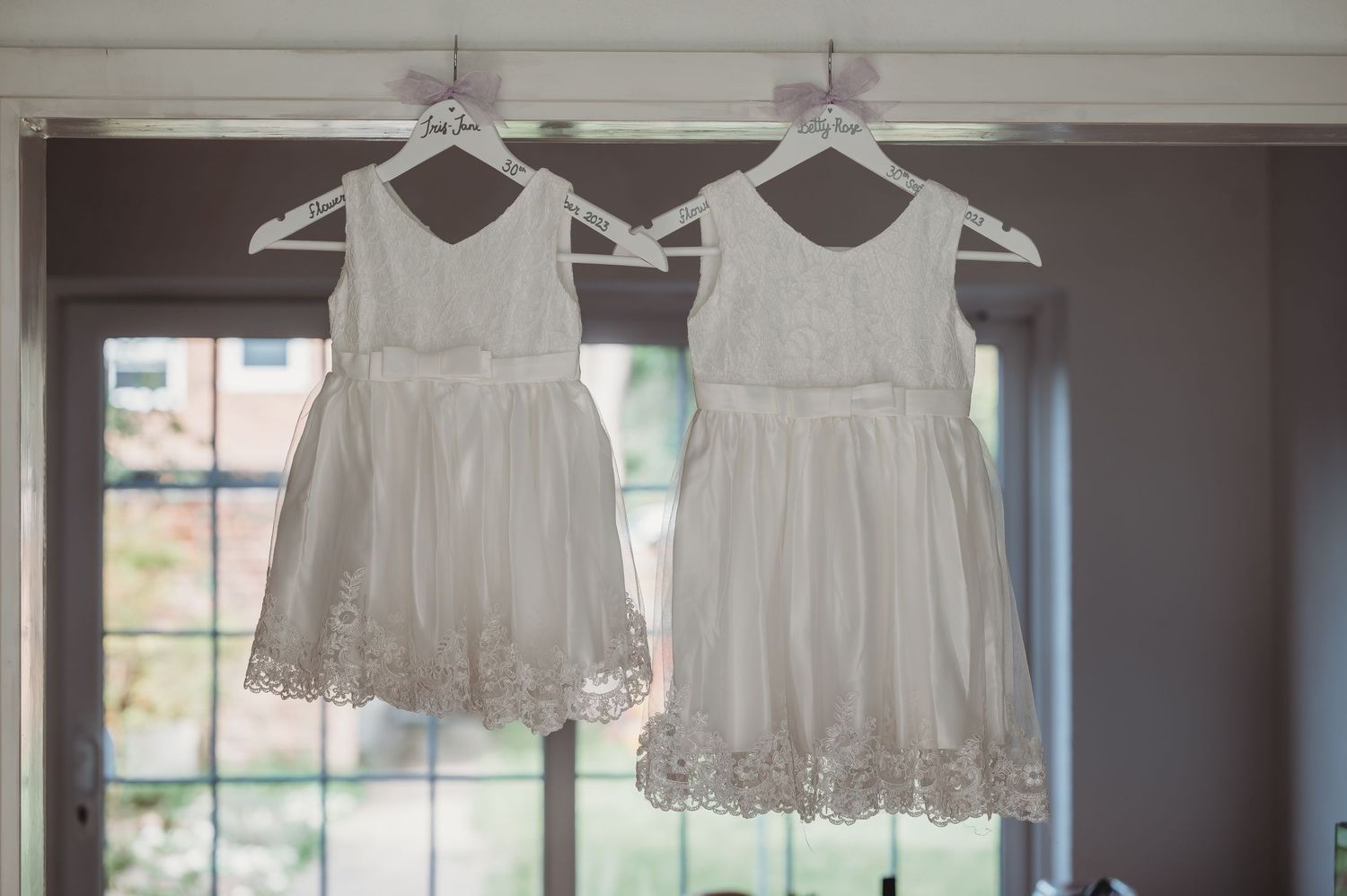 Two delicate white flower girl dresses with lace trim hanging side by side from hangers in a window.
