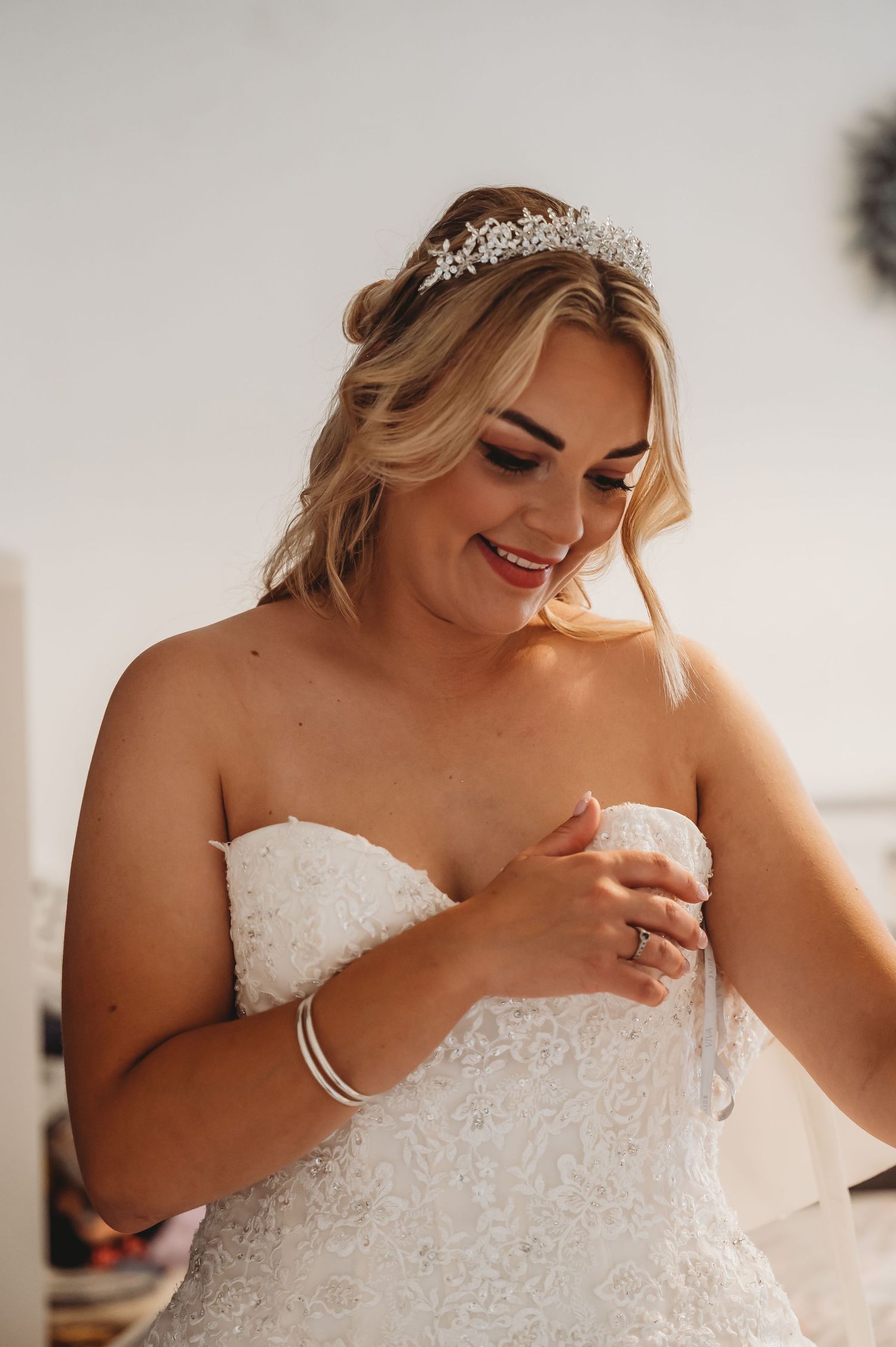 A bride wearing a strapless lace wedding gown and crystal tiara smiles while getting ready for her special day.