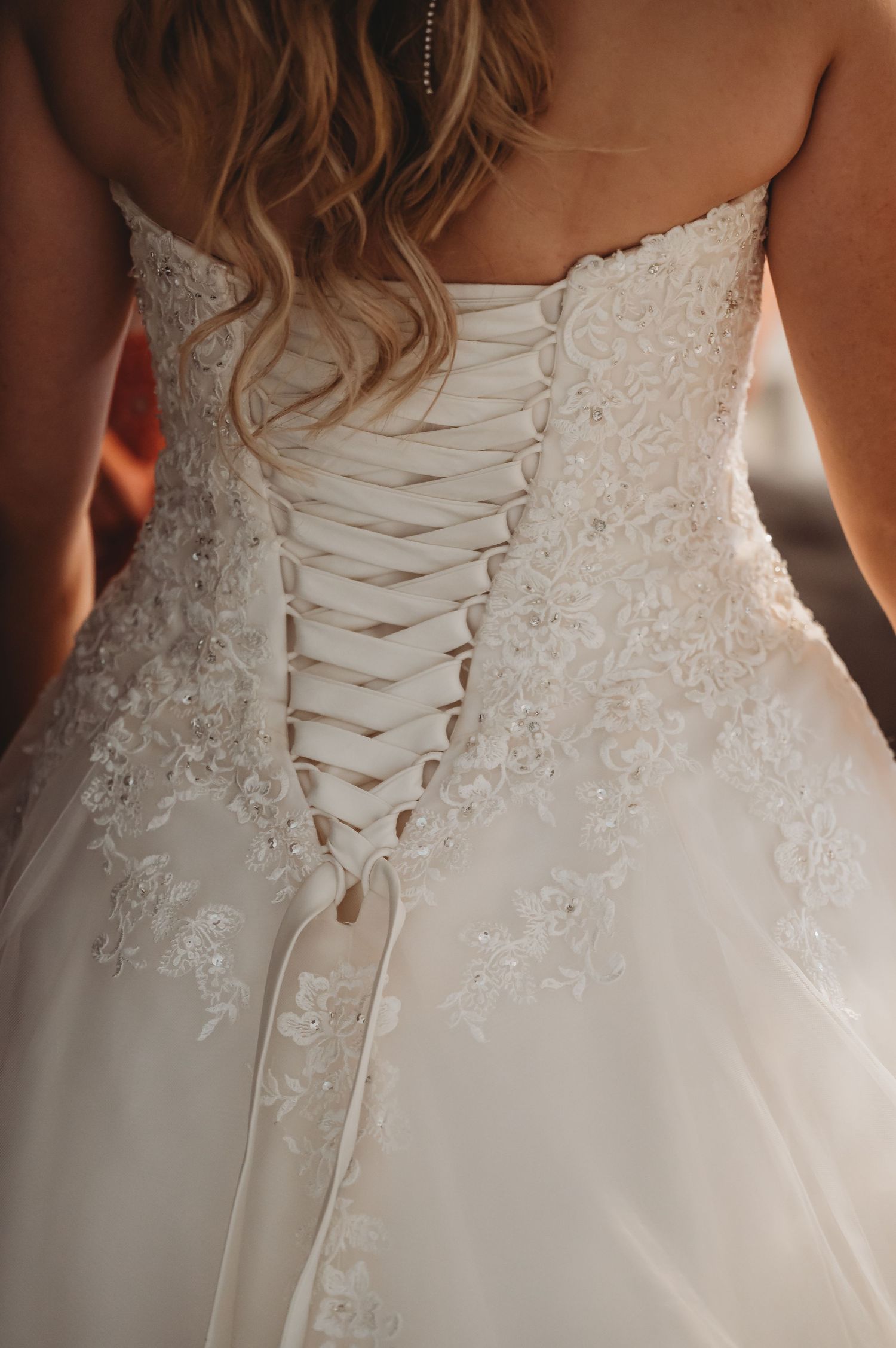 Close-up detail of a white wedding dress showing intricate lace pattern and corset-style ribbon lacing up the back.