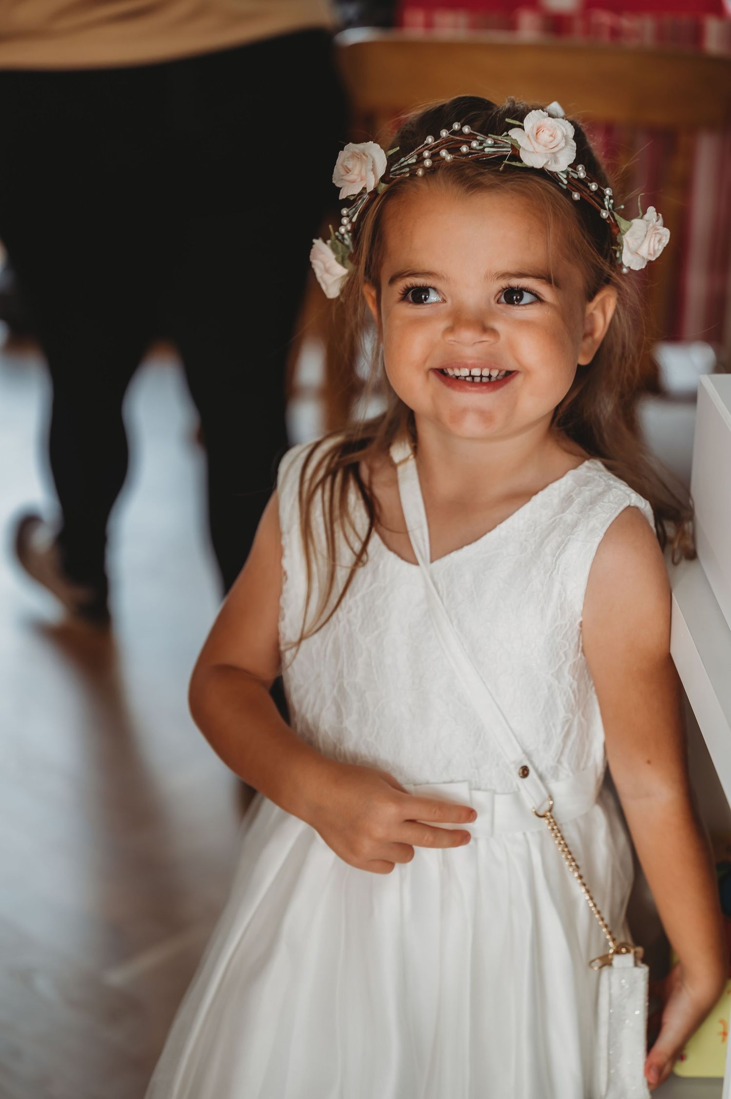 Toddler wearing white flower girl dress and floral crown smiles during wedding celebration.