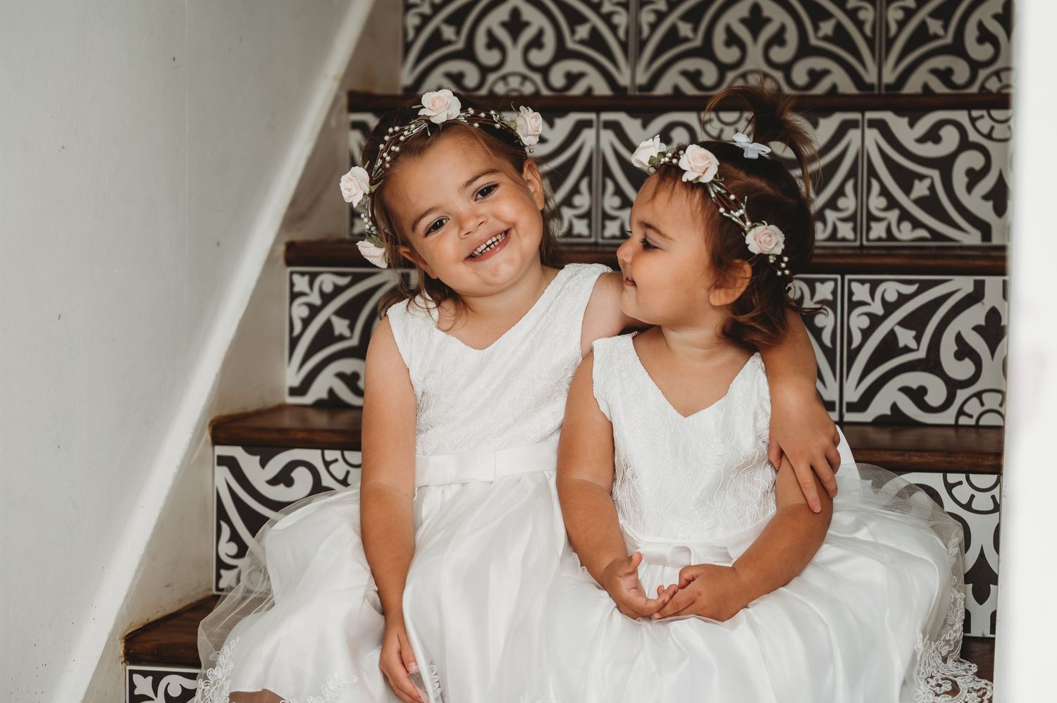 Two flower girls in white dresses laugh together while sitting on decorative stairs at a wedding.
