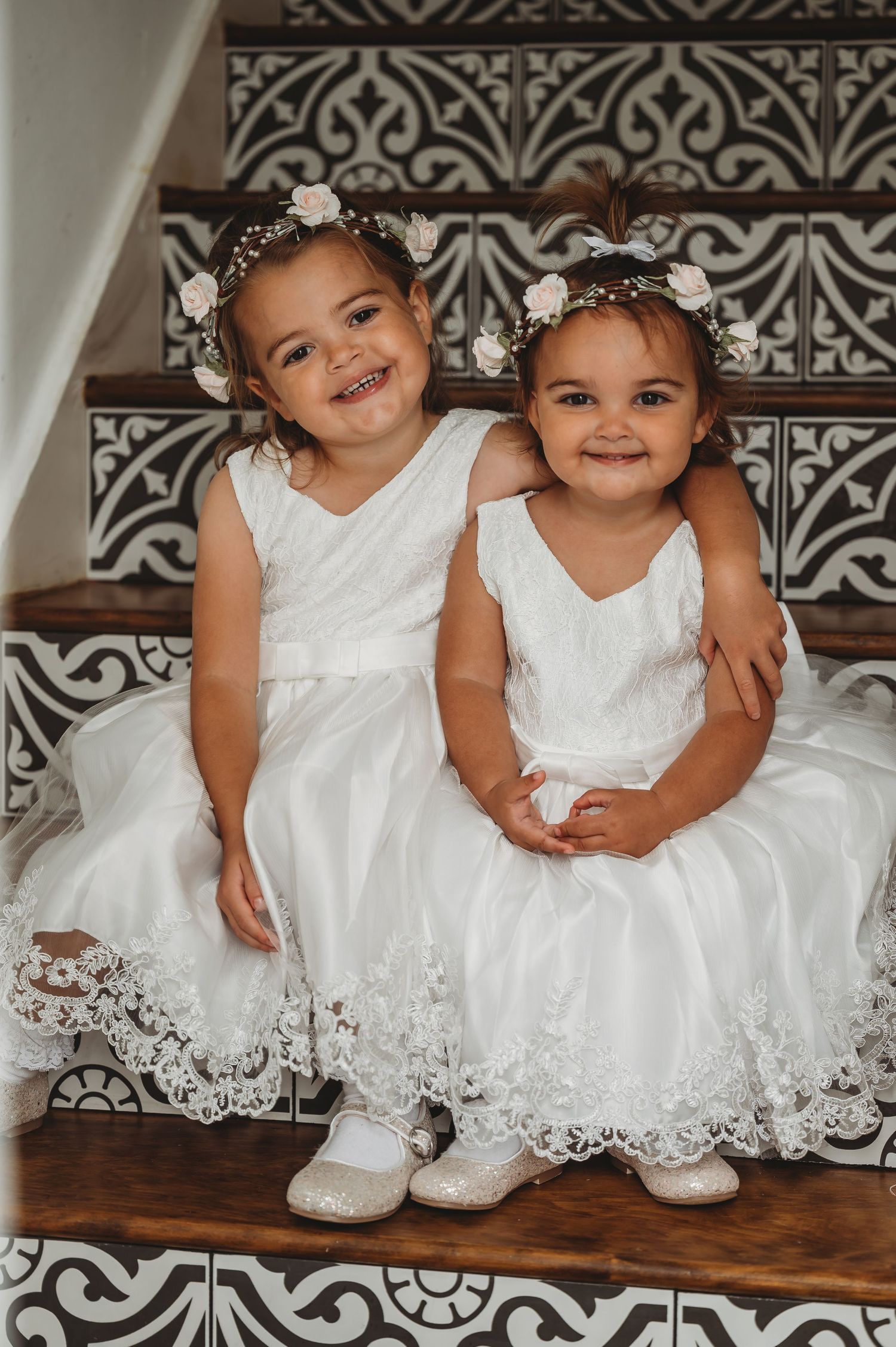 Two young children in matching white lace dresses and floral headpieces sit together on decorative stairs.