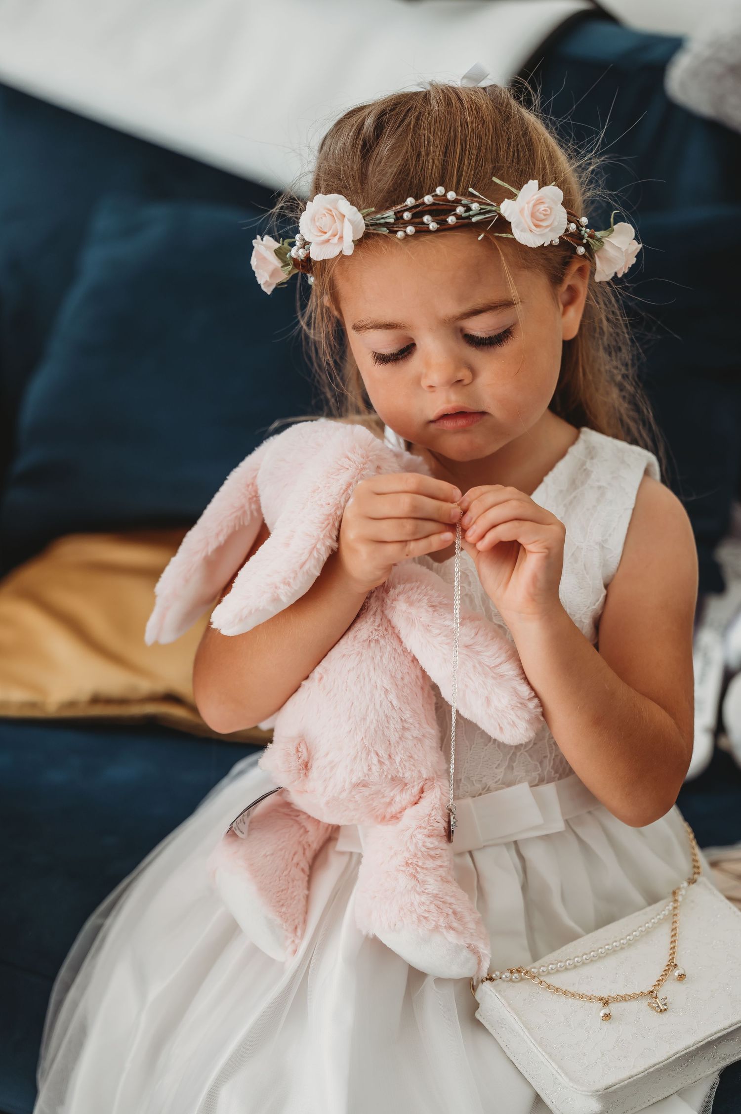 A young child in a white dress wears a floral crown and holds a pink plush toy while sitting on a blue couch.