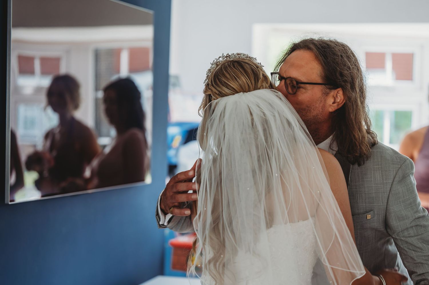 A bride in a wedding veil is embraced by a person with long hair and glasses at an indoor venue.