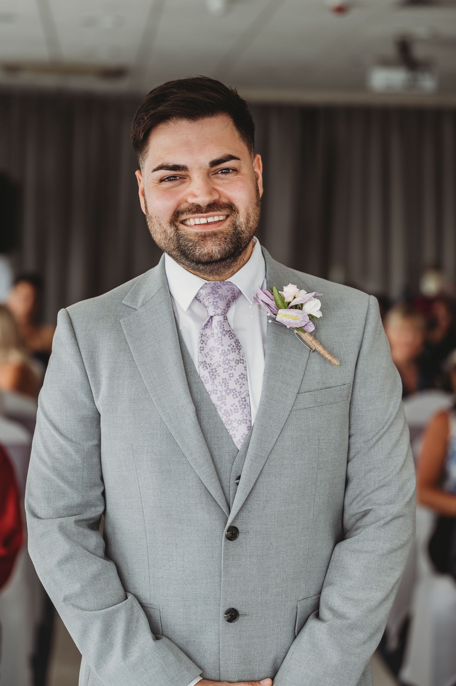 A groom in a light grey suit with pink tie and floral boutonniere smiles warmly at his wedding.