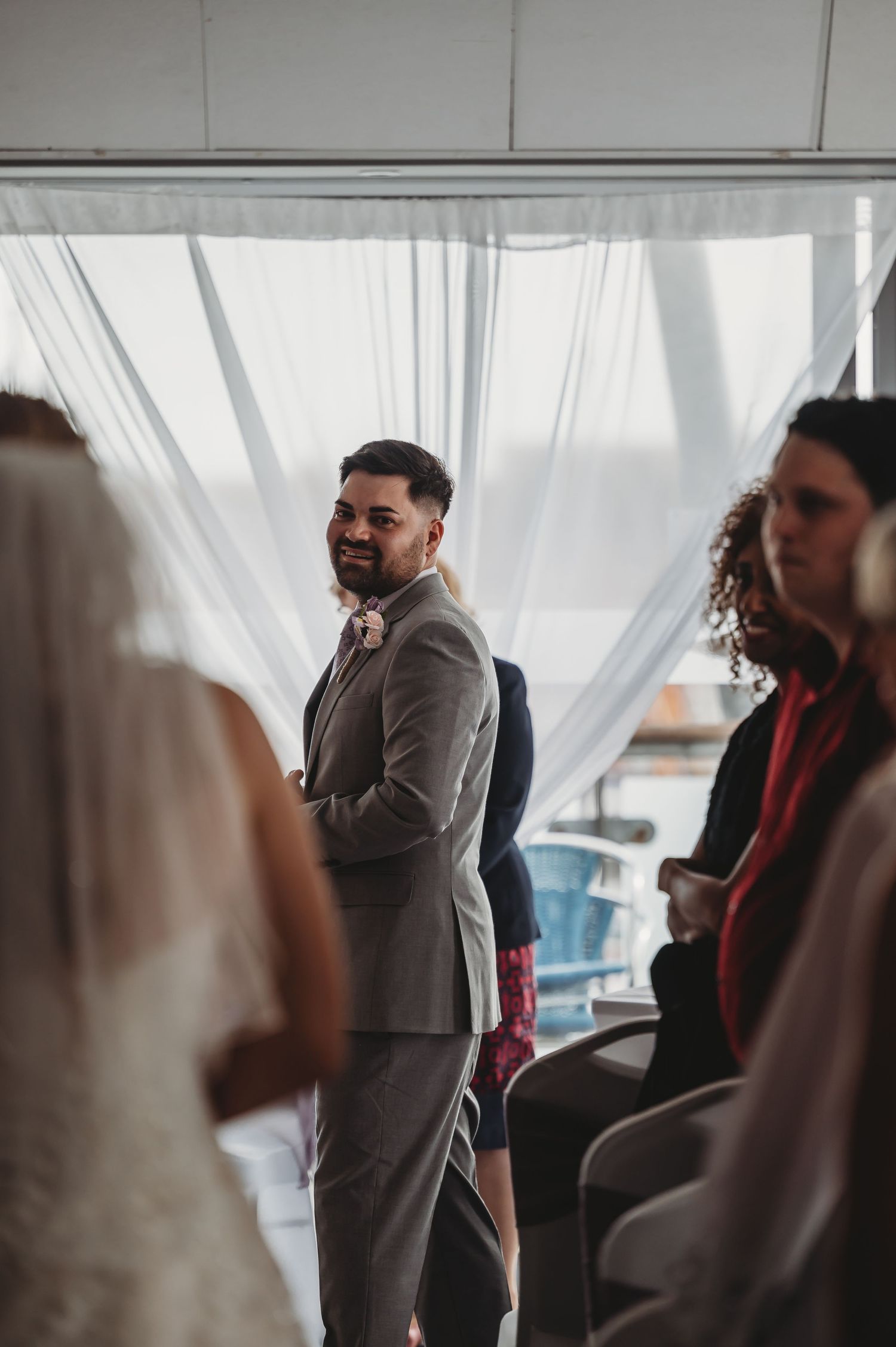 A groom in a grey suit stands at attention during a wedding ceremony, with guests visible in soft focus.