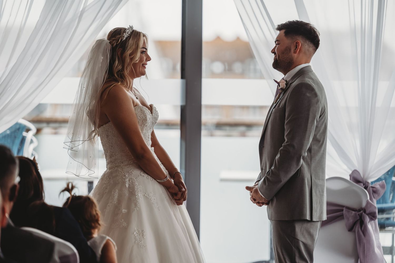 A couple exchanges vows during their wedding ceremony at a venue with flowing white curtains and waterfront views.
