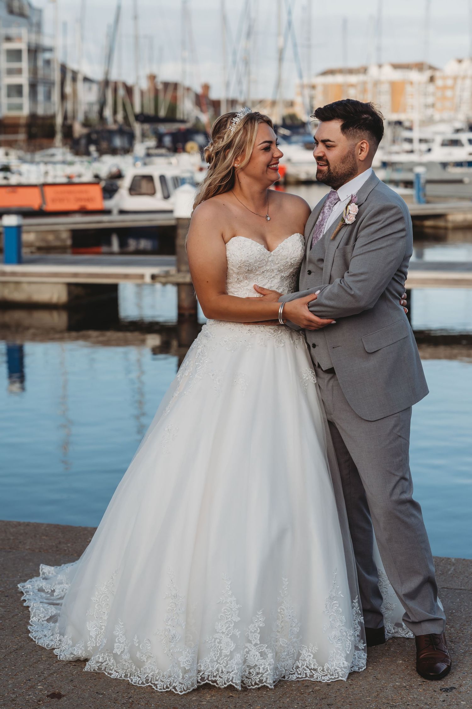 A romantic newlywed couple embraces at a marina with sailboats and modern buildings in the background.