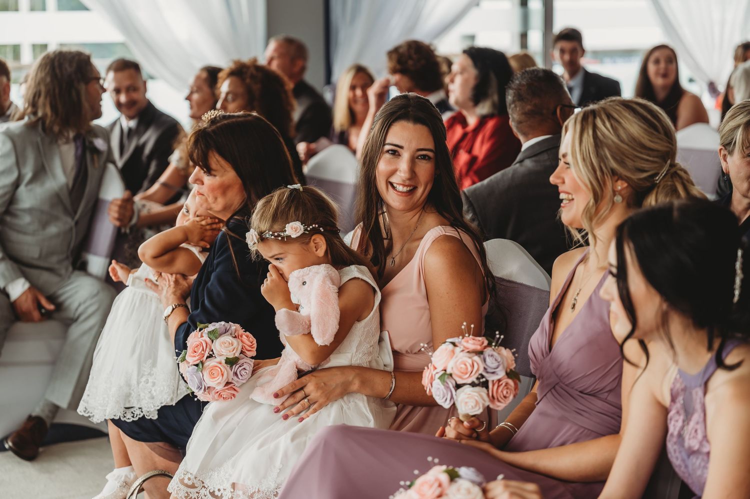 Wedding party members seated in a row holding pink bouquets during ceremony with guests visible in background.