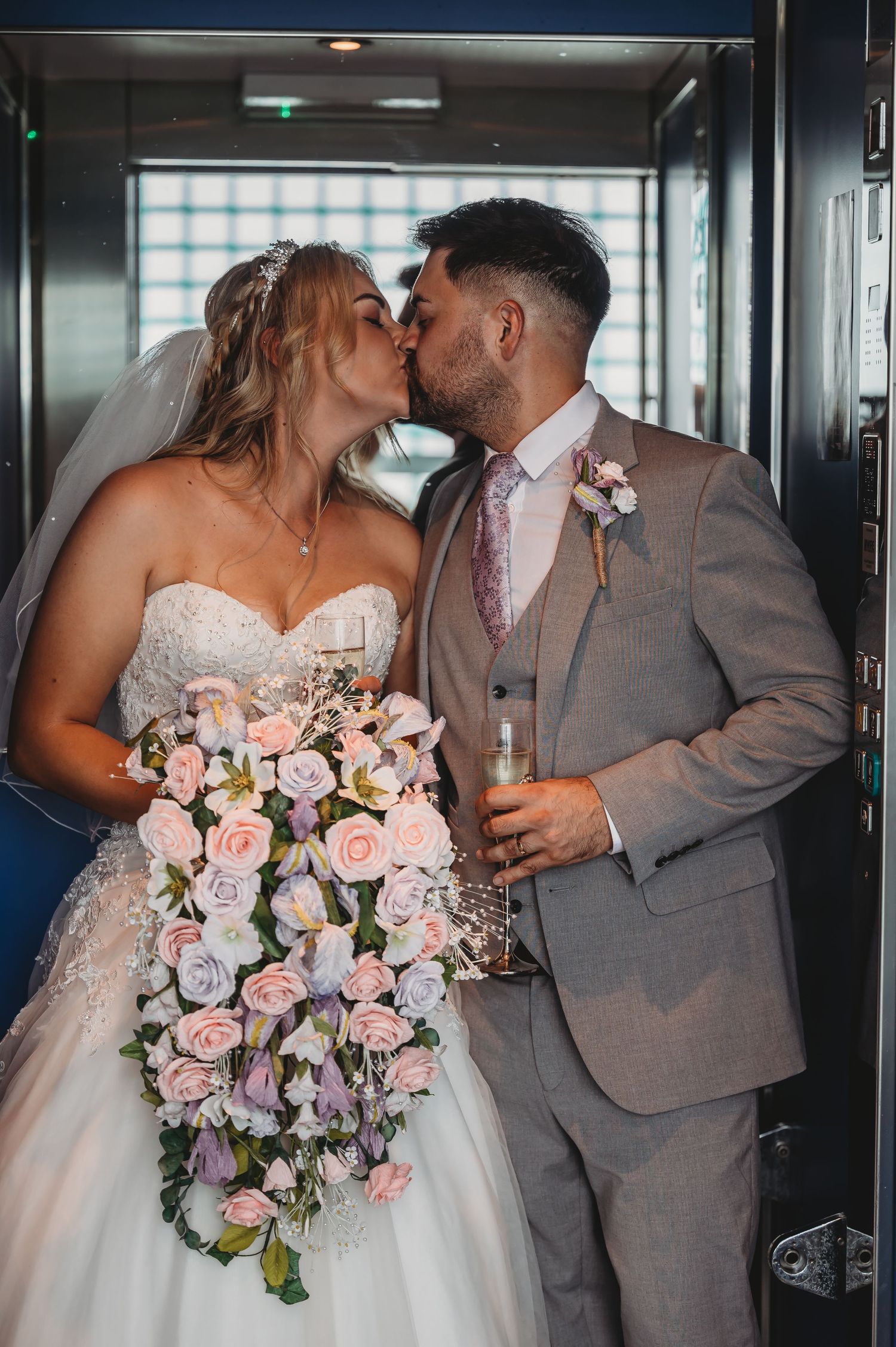 A couple shares a wedding day kiss in an elevator, with the bride holding a cascading bouquet of pink roses.
