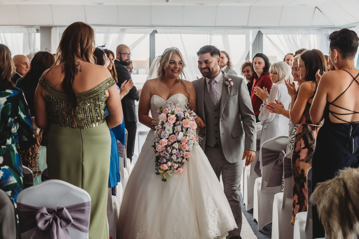 Wedding couple smiles joyfully while walking down aisle surrounded by guests at indoor ceremony.