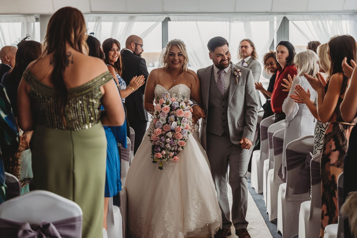A wedding ceremony processional in a light-filled venue with guests looking on.