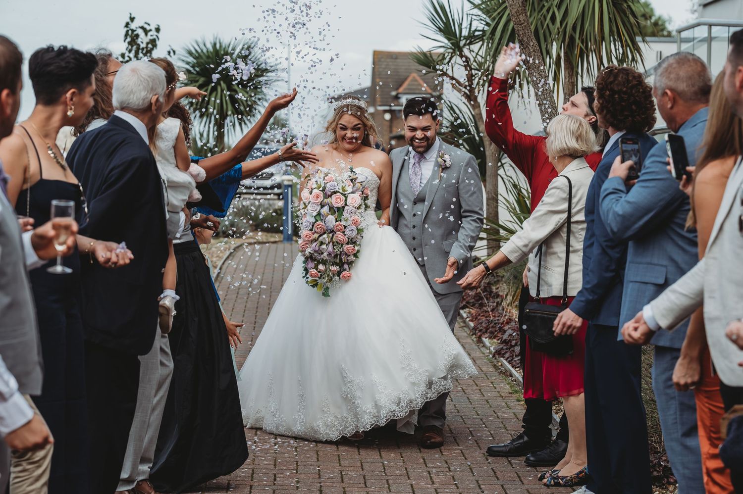 Wedding guests joyfully toss confetti over newlyweds walking down brick pathway at outdoor celebration.