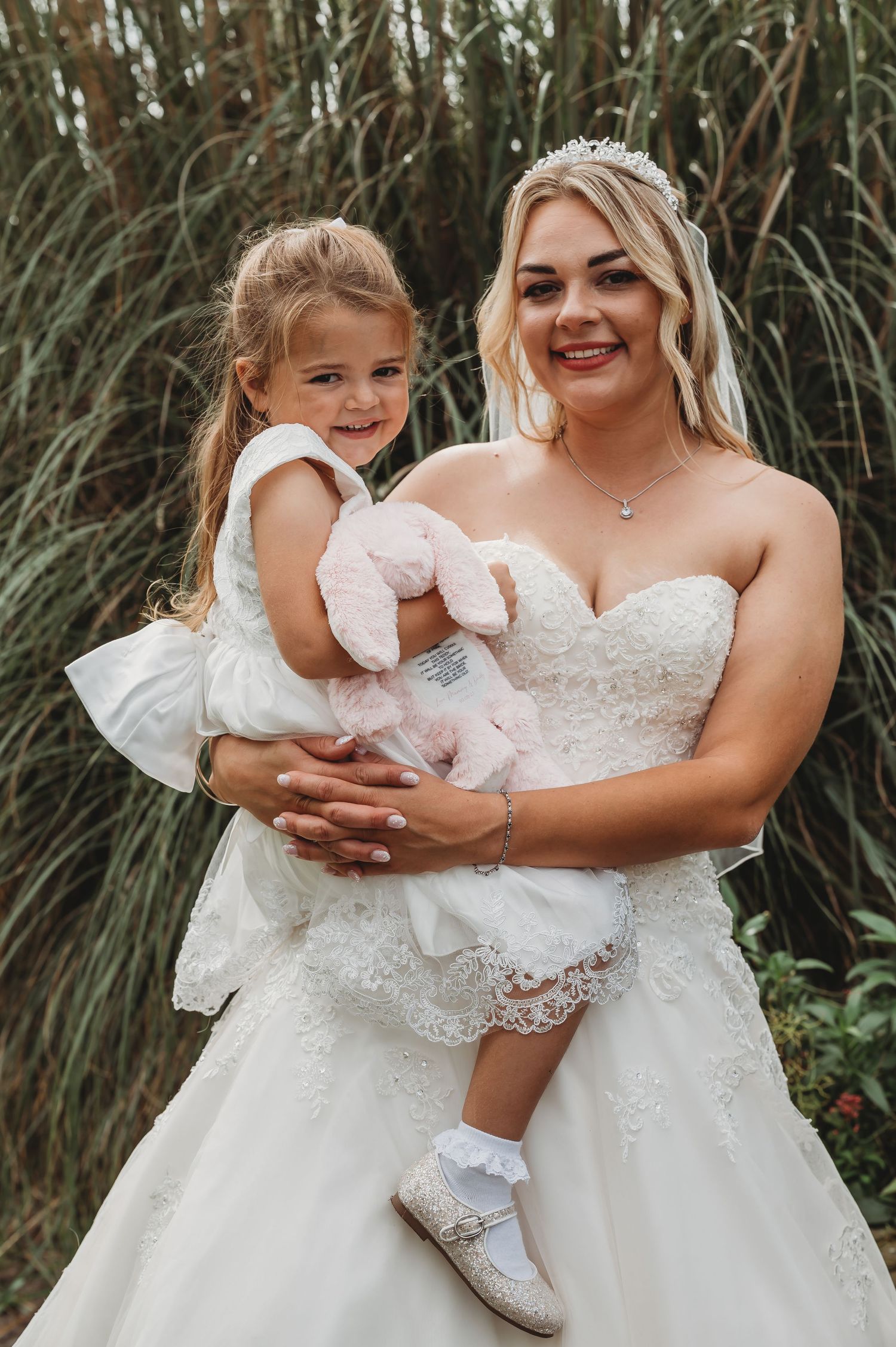 Beautiful bride in white lace dress holds young flower girl in matching white dress among tall reeds outdoors.
