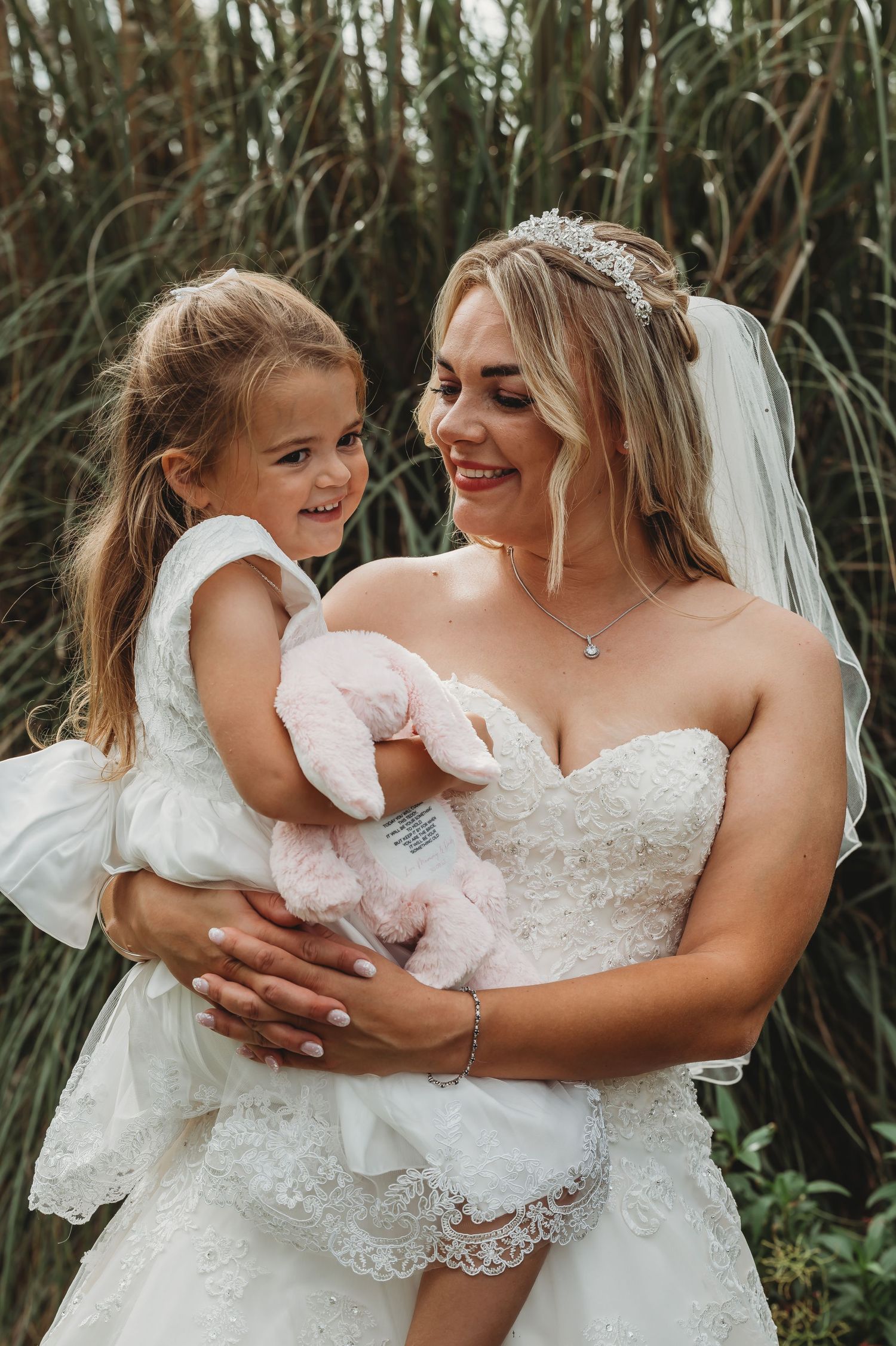 A heartwarming moment captured outdoors in tall grass as a bride in a lace gown holds a young flower girl wearing white.