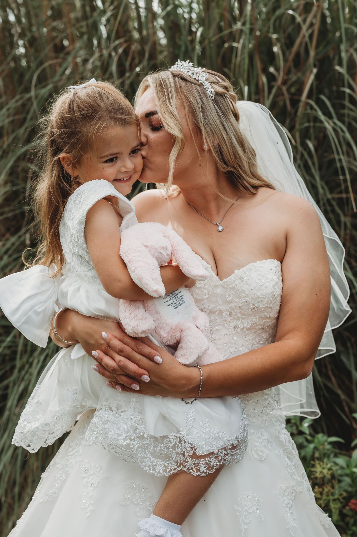 Sweet moment between a bride in a white lace wedding dress and a young flower girl amid tall reeds.