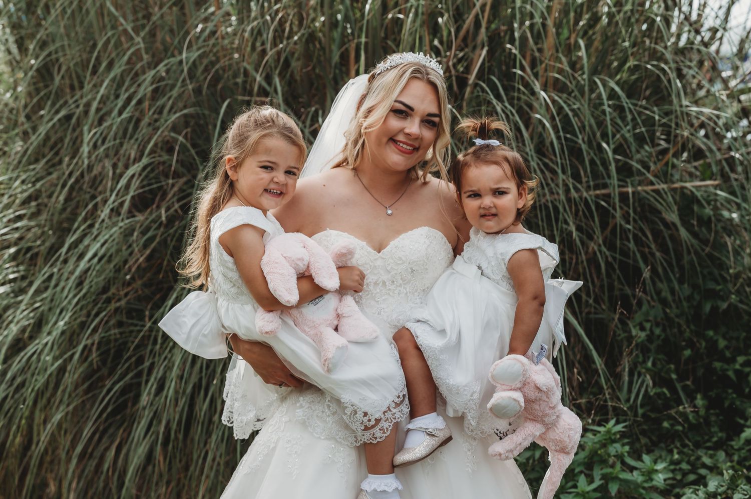 A bride in a white wedding dress holds two young children wearing white dresses against a backdrop of tall grass.