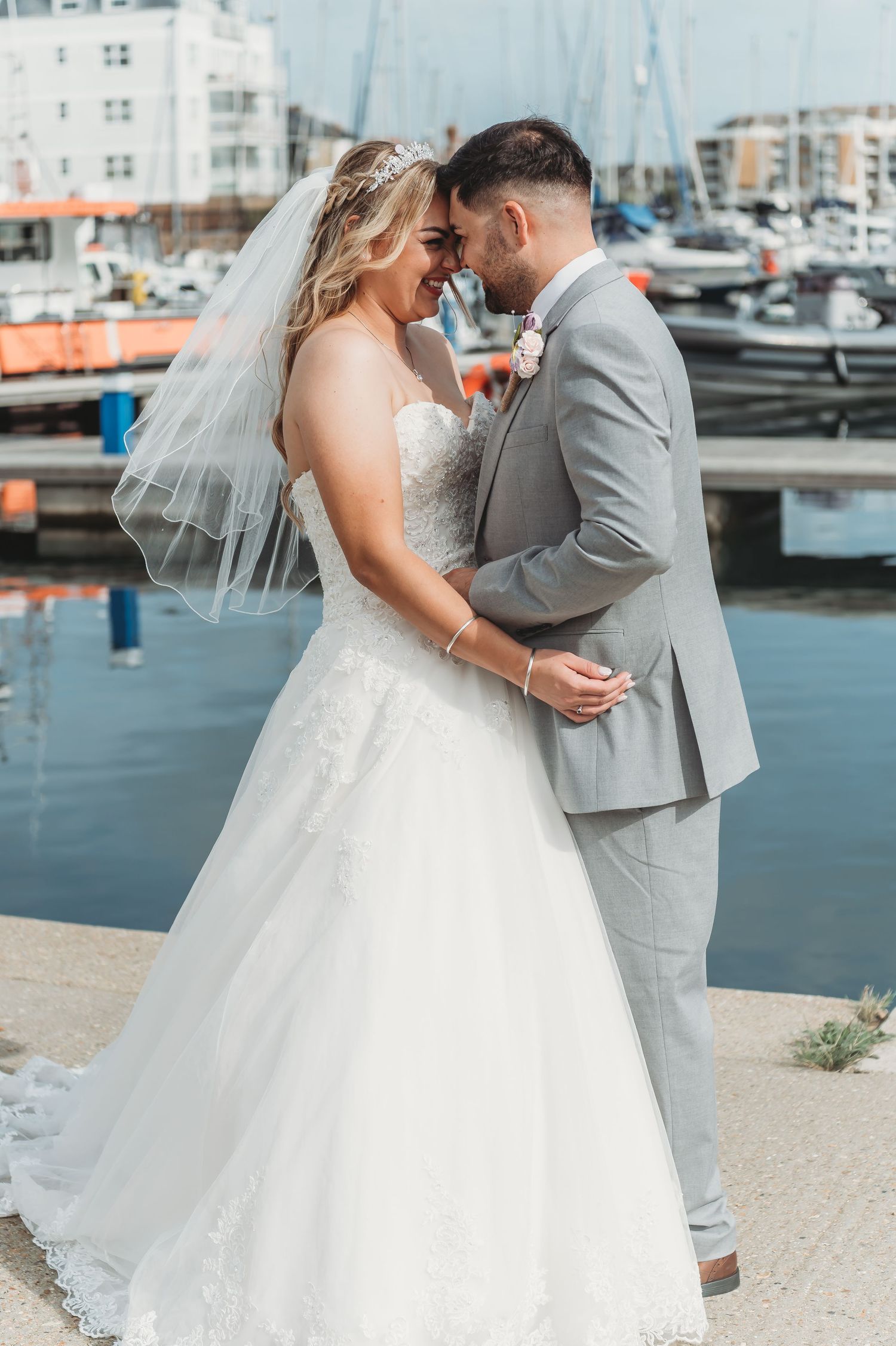 A newlywed couple shares an intimate moment by a marina with boats and buildings in the background.