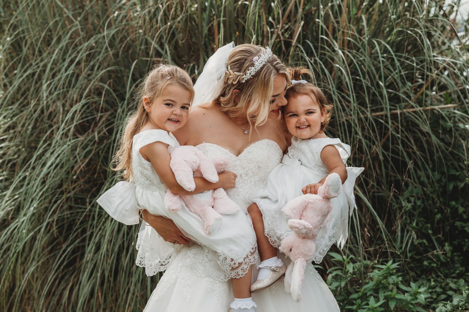 A bride in a white wedding dress holding two little ones in matching white outfits against tall ornamental grass.