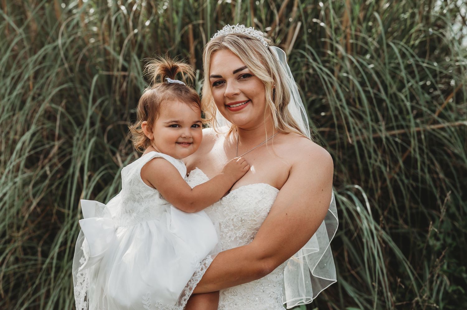 Mother and daughter in white dresses share a loving embrace against tall grass background.