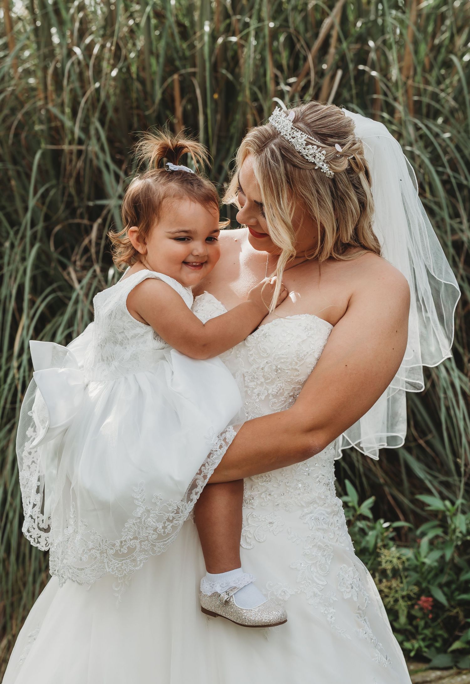 A tender moment between a bride in a white lace dress and a small girl in a white dress against tall reeds.
