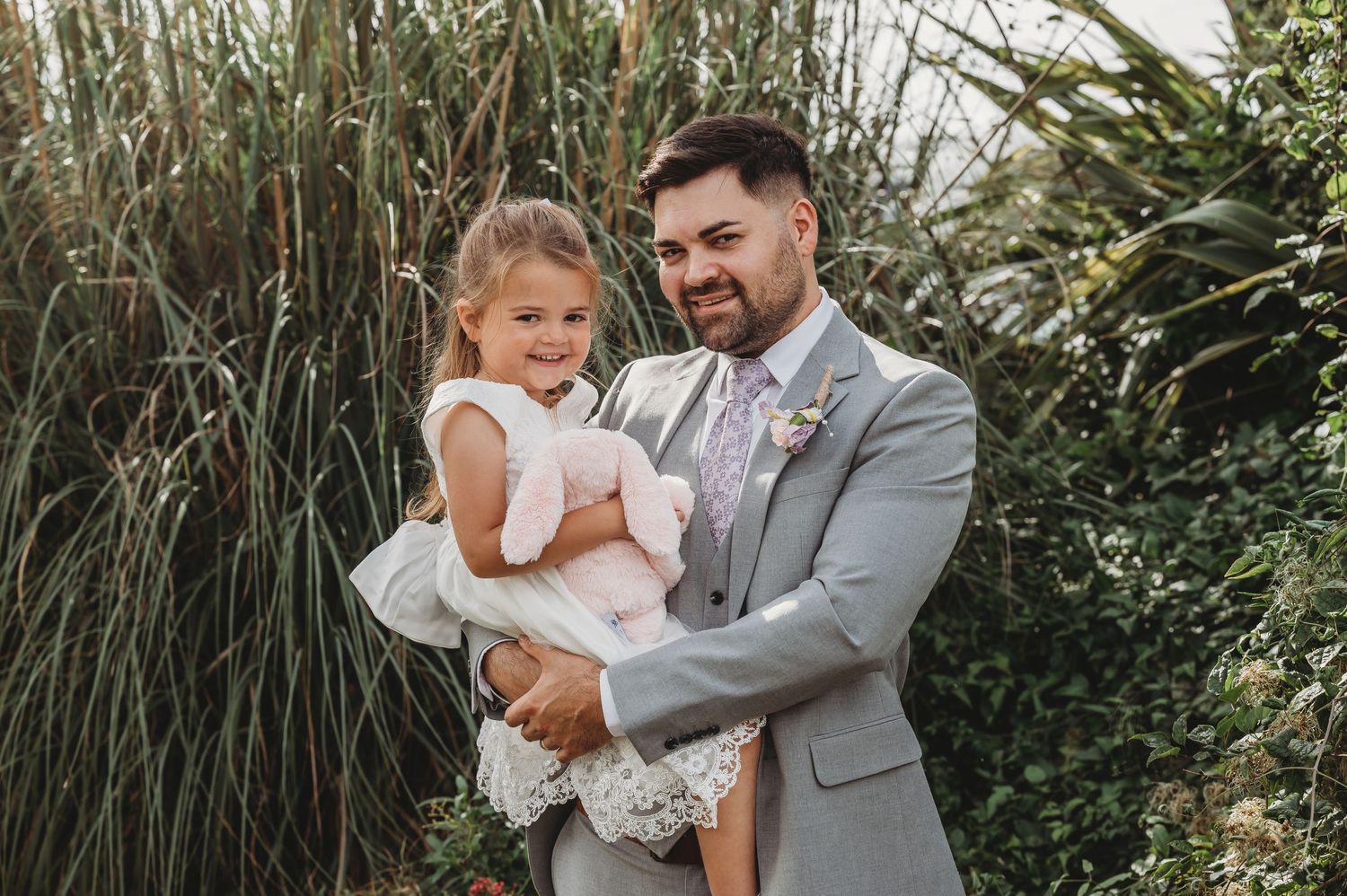A parent wearing a gray suit holds a child in a white dress among tall ornamental grasses in an outdoor setting.