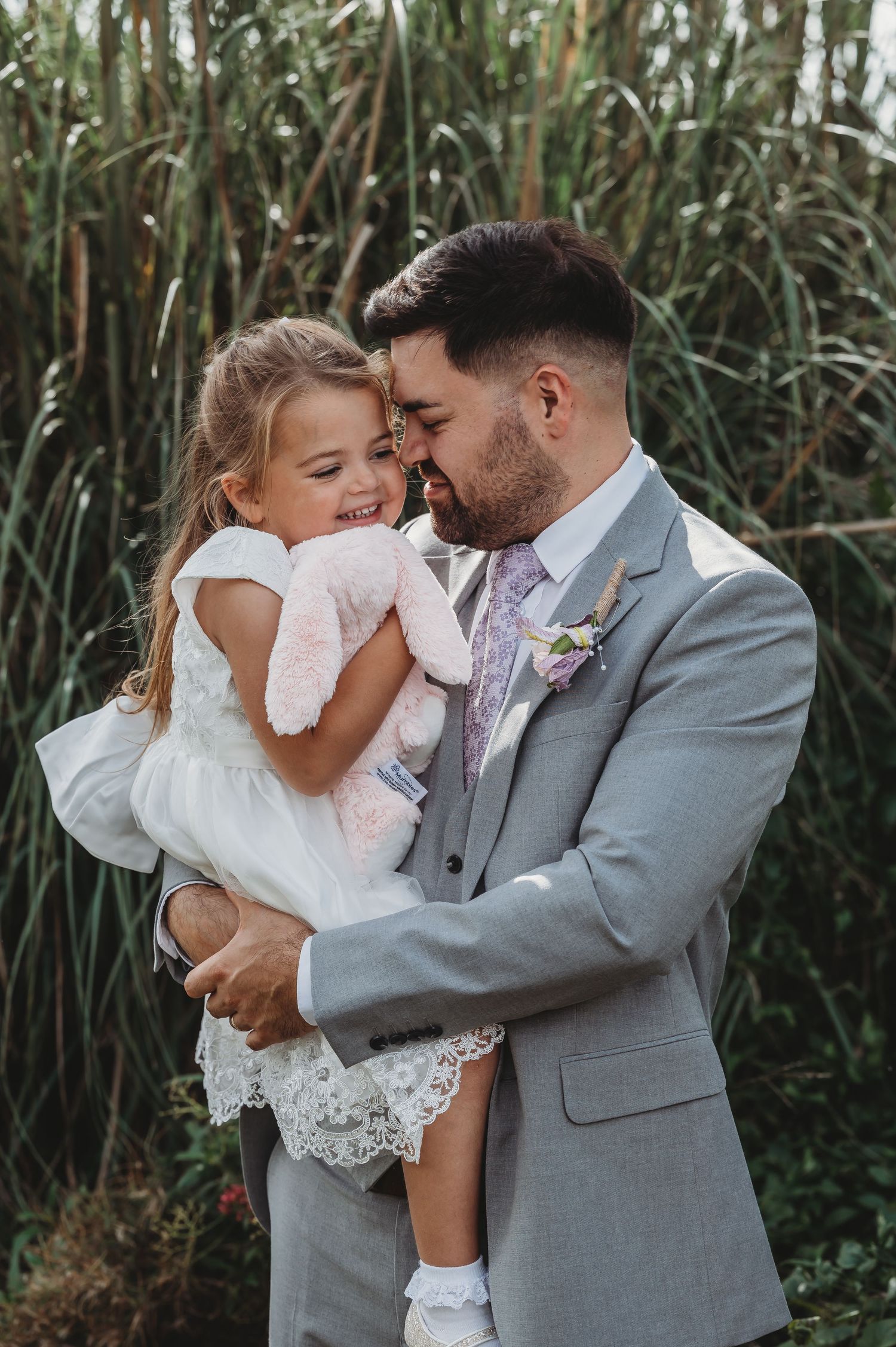 Touching candid wedding moment shared between groom in gray suit and young girl in white dress against tall grass backdrop.