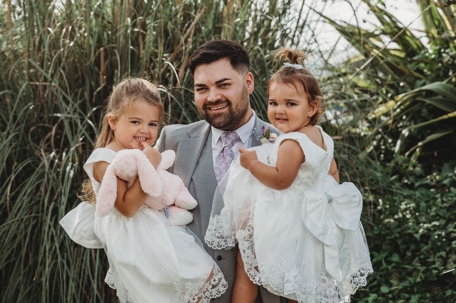 A joyful scene in a garden shows three people in white attire smiling together against tall ornamental grasses.