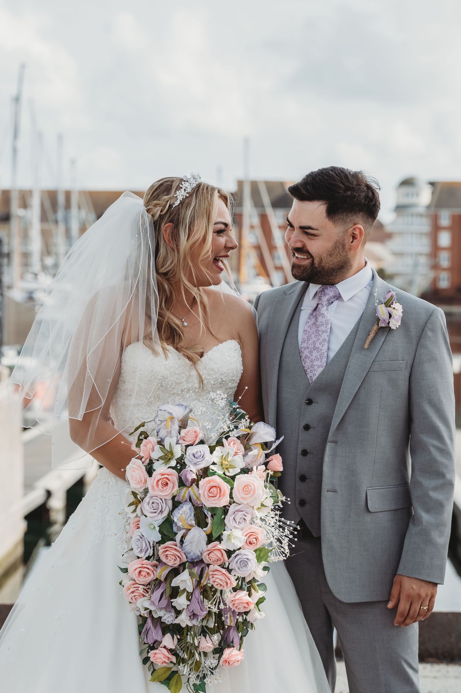 A bride in a white dress holding a pastel pink and purple floral bouquet stands with a groom in a gray suit at a marina.