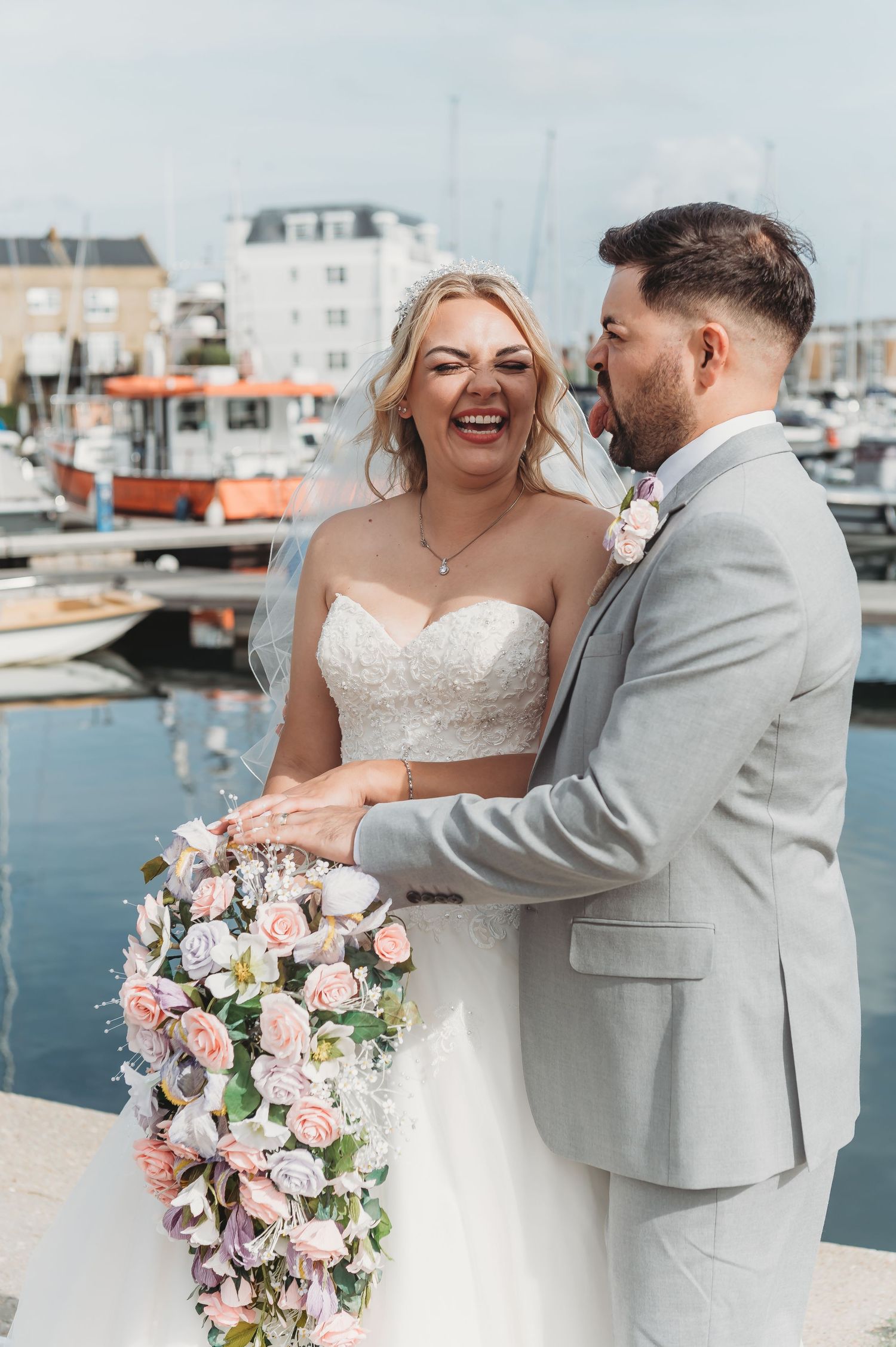 A joyful moment shared by a couple in wedding attire holding a cascading pink and white floral bouquet by a marina.