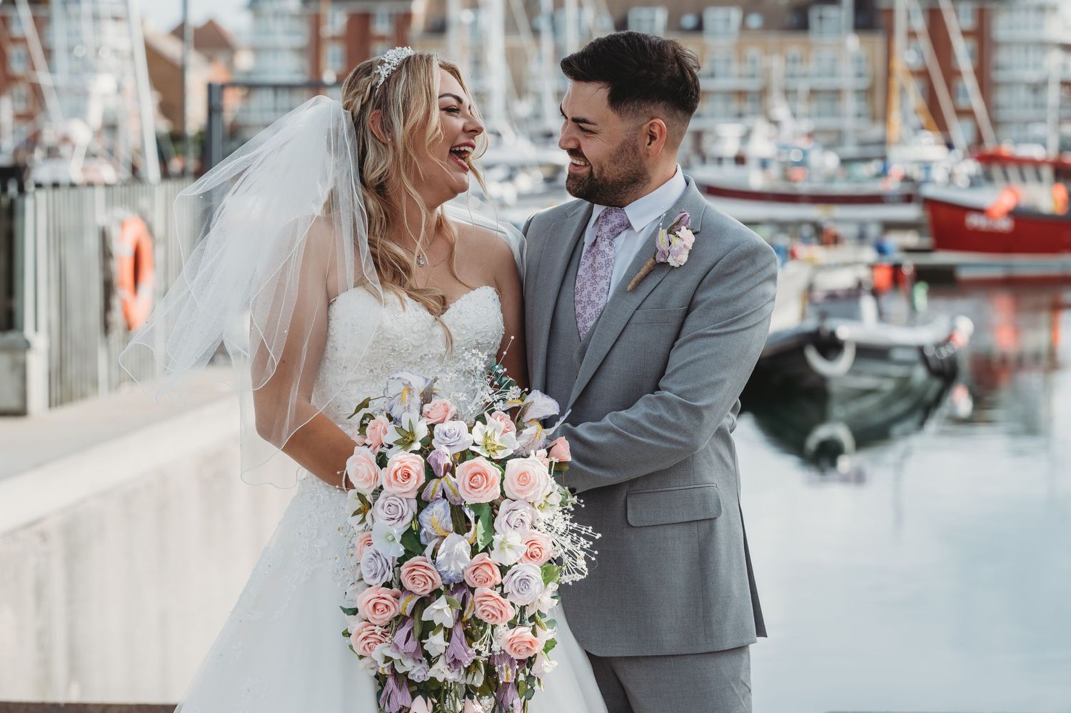 A newlywed couple shares an intimate moment at a marina with boats in background, bride holding pink and white bouquet.