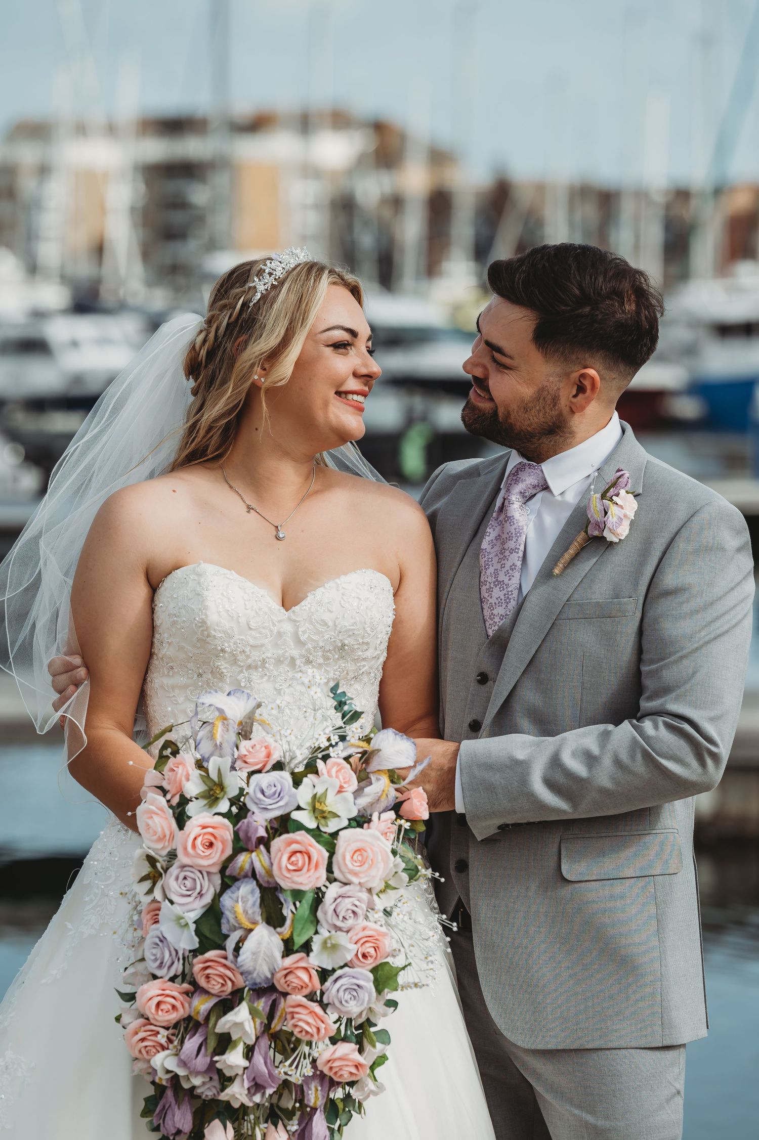 A newlywed couple shares a joyful moment at a marina with boats in the background, showcasing a beautiful floral bouquet.